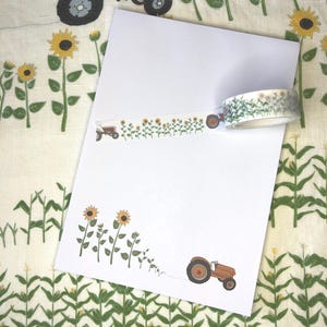 Könnte beinhalten: Eine Rolle dekoratives Washi-Tape mit einem sich wiederholenden Muster aus kleinen braunen Traktoren und gelben Sonnenblumen. Ein Streifen des Tapes ist auf einem weißen Blatt Papier angebracht und zeigt dasselbe Motiv in einer horizontalen Linie.