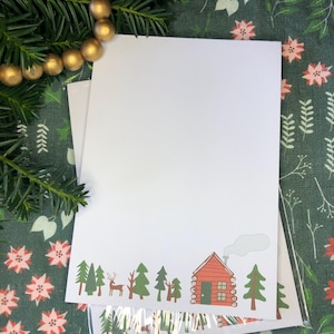 May include: White stationery with a cabin and forest design. The illustration features a red cabin with a smoking chimney, green trees, and a deer. The stationery is packaged in a clear plastic sleeve.