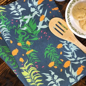 May include: A navy blue cloth napkin with a botanical pattern featuring green, orange, and light blue leaves and flowers. A wooden spoon, a plate with cookies, and a cup of coffee are also visible.
