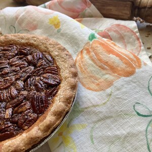Peut inclure: Une tarte aux noix de pécan avec une croûte dorée et une garniture sombre et brillante recouverte de noix entières, posée sur un tissu blanc orné d'un motif aquarelle de citrouilles orange et de vignes vertes, avec un rouleau à pâtisserie en bois.