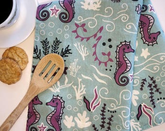 Puede incluir: Un paño de cocina azul claro con un patrón repetitivo de caballitos de mar morados, coral blanco, algas de color marrón oscuro y líneas onduladas. La tela está decorada con pequeños círculos blancos y estrellas en todo el diseño acuático.