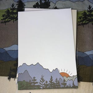Könnte beinhalten: Ein Stapel leerer weißer Karten mit einem Landschaftsdesign. Das Design zeigt blaue Berge, grüne Bäume und einen orangefarbenen Sonnenuntergang. Die Karten liegen auf einem gemusterten Stoff mit einem ähnlichen Berg- und Baumdesign.
