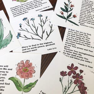 Könnte beinhalten: Eine Sammlung von Aquarell-Blumenkunstkarten. Jede Karte zeigt eine einzigartige Blumenillustration in Rosa-, Rot- und Blautönen, begleitet von biblischem Text. Die Karten sind auf einer dunklen Holzoberfläche angeordnet.