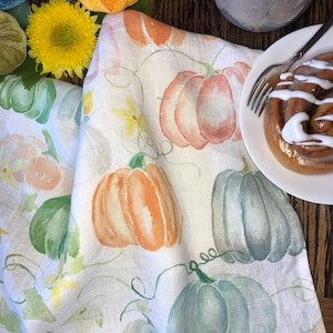 May include: A white kitchen towel featuring a watercolor print of pumpkins in shades of orange, sage green, and muted blue, accented with thin green vines and small yellow flowers, arranged in a repeating pattern across the fabric surface.