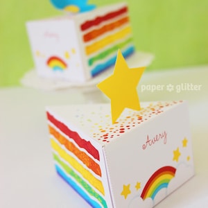 Könnte beinhalten: Eine regenbogenförmige Kuchenbox mit weißem Hintergrund und einem gelben Stern oben. Die Box hat ein Regenbogenmuster an der Seite und den Namen "Avery" auf der Vorderseite. Die Box ist mit kleinen gelben Sternen und einem Regenbogen verziert.