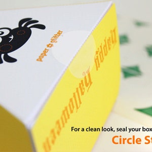 May include: A yellow and white triangular box with a black spider illustration and the text "paper & glitter" and "happy halloween". The text "For a clean look, seal your boxes with clear Circle Stickers" is at the bottom of the image.