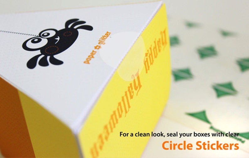 May include: A yellow and white triangular box with a black spider illustration and the text "paper & glitter" and "happy halloween". The text "For a clean look, seal your boxes with clear Circle Stickers" is at the bottom of the image.
