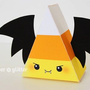 May include: A yellow, orange, and white candy corn shaped box with black bat wings. The box has a cute cartoon face with fangs and rosy cheeks. The text "paper * glitter" is printed on the bottom of the box.