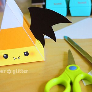 May include: A paper craft project of a candy corn shaped box with a black bat wing design. The box has a cute cartoon face with fangs. The image shows the box, scissors, and a craft knife on a wooden surface.
