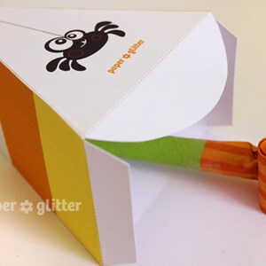 May include: A white paper party favor box with orange, yellow, and white stripes. The box is open and a green and orange party blower is inside. The box has a spider design and the text "paper & glitter" on the front.