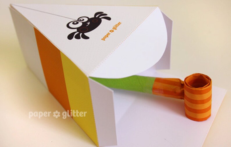 May include: A white paper party favor box with orange, yellow, and white stripes. The box is open and a green and orange party blower is inside. The box has a spider design and the text "paper & glitter" on the front.