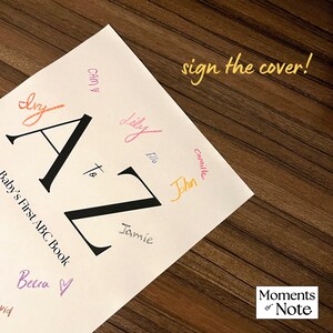 May include: A white "Baby's First ABC Book" with large black "A to Z" letters. The cover is signed with colorful names. Text reads "sign the cover!" and "Moments of Note" on a wooden surface.