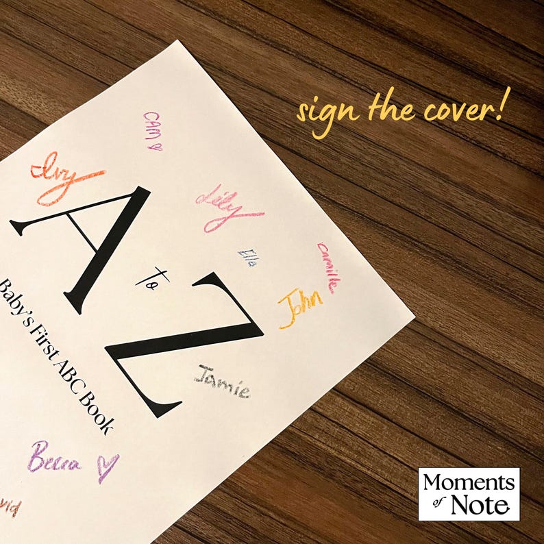 May include: A white "Baby's First ABC Book" with large black "A to Z" letters. The cover is signed with colorful names. Text reads "sign the cover!" and "Moments of Note" on a wooden surface.