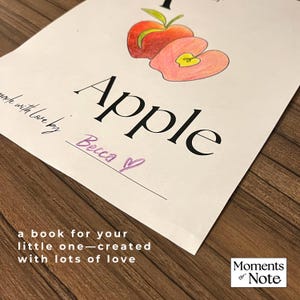 May include: A hand-drawn illustration of an apple with the word "Apple" in large black letters. The text "made with love by Becca" and "a book for your little one - created with lots of love" are also present. The image is on a wooden surface.