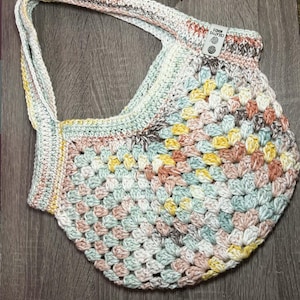 May include: A handmade crochet bag in a granny square pattern. The bag features a mix of pastel colors including yellow, pink, blue, and cream. The bag has a single strap and a small tag that reads "CREATIVE MINDS".