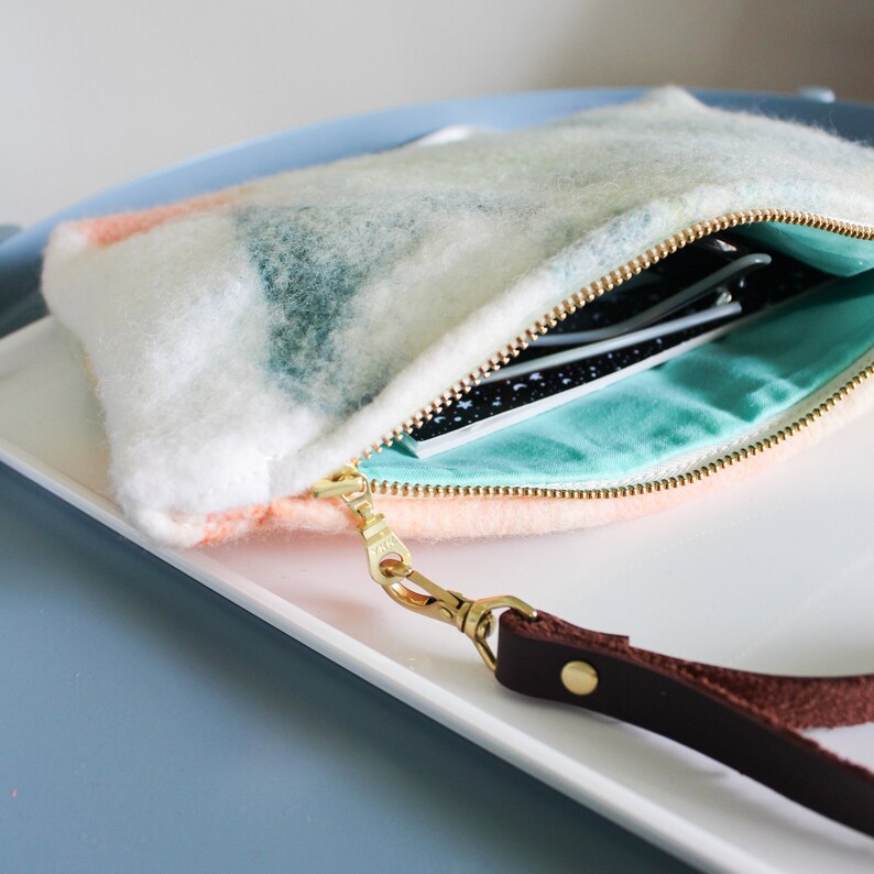 May include: A white, pink, and green fuzzy clutch with a gold zipper and a brown leather strap. The clutch is open and has a turquoise lining.