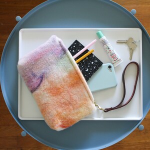 May include: A colorful, fuzzy pouch with a zipper closure and a brown leather strap. The pouch is filled with a notebook, pencils, a tube of lip balm, and a set of keys.