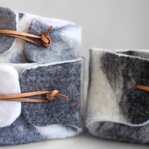 May include: Three gray and white felt storage baskets with brown leather straps. The baskets are stacked on top of each other.