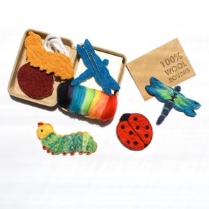 May include: A craft kit containing felted wool shapes including a green caterpillar, a red ladybug, and a blue dragonfly. It includes a rainbow-colored wool roving roll, a brown cardboard container, and a card labeled 100% WOOL ROVING.