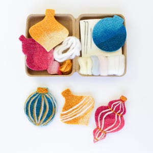 May include: A craft kit with felt ornaments in shades of orange, pink, and blue. The kit includes felt shapes, white yarn, needles, and other materials. Three finished ornaments are also displayed, showcasing the design possibilities.