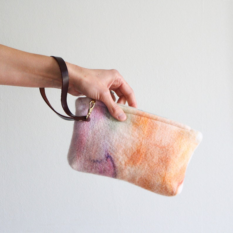 May include: A soft, pastel-colored clutch bag with a brown leather strap. The bag has a subtle tie-dye pattern in shades of pink, orange, and yellow.