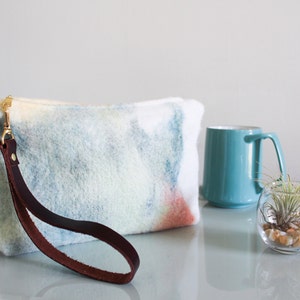 May include: A light blue and white tie-dye clutch with a brown leather strap. The clutch is made of a soft, fuzzy material and has a gold clasp.