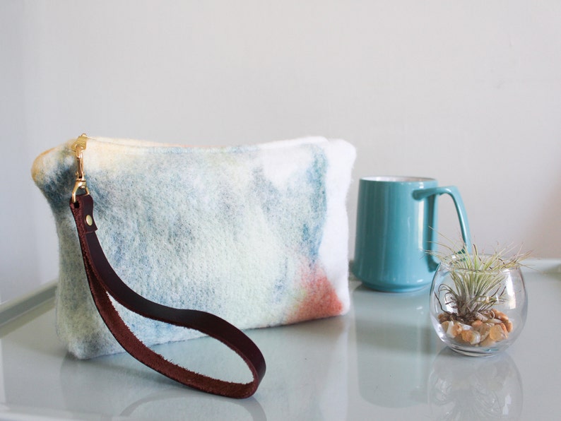 May include: A light blue and white tie-dye clutch with a brown leather strap. The clutch is made of a soft, fuzzy material and has a gold clasp.