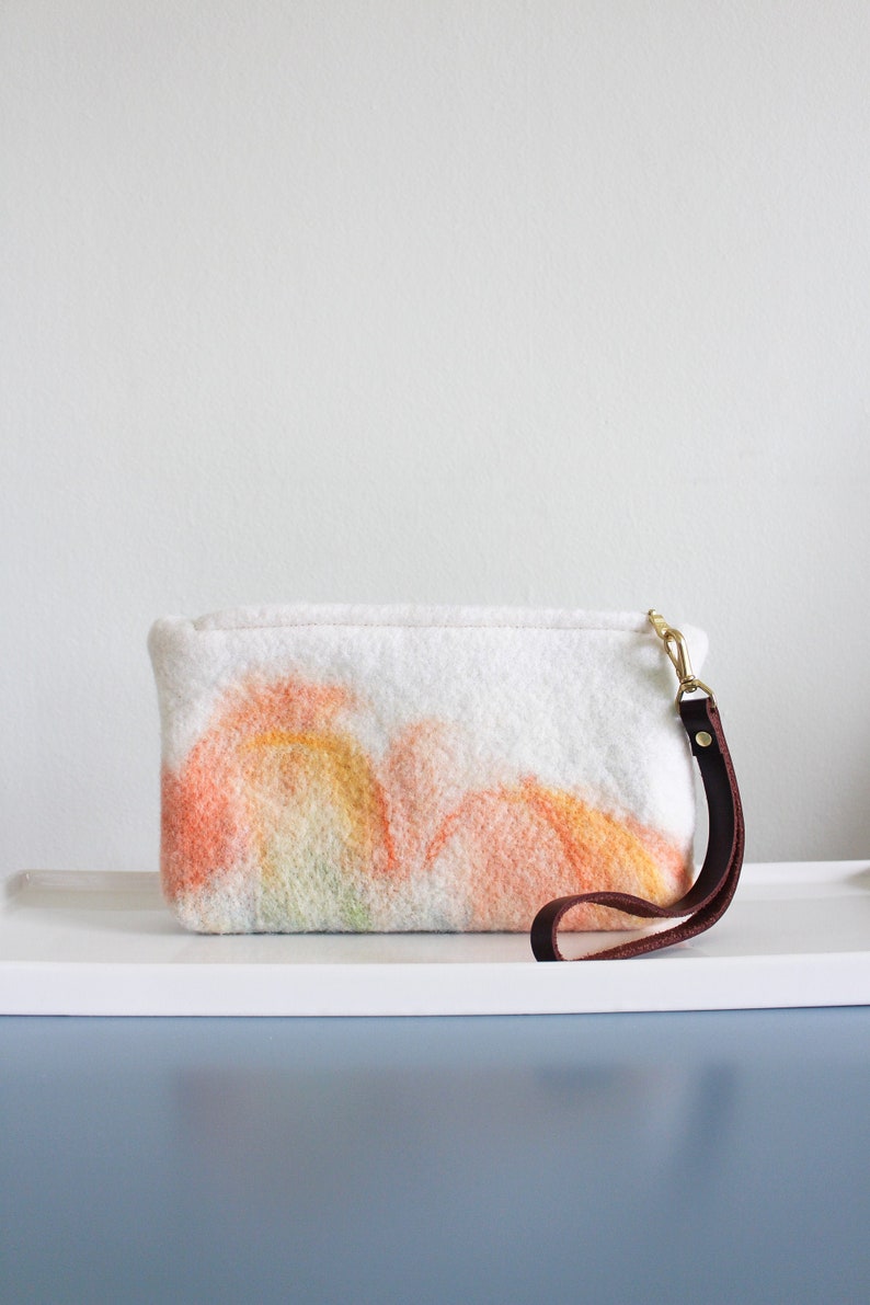 May include: A white felt clutch bag with a brown leather strap and a colorful abstract design. The design features shades of orange, yellow, and green.