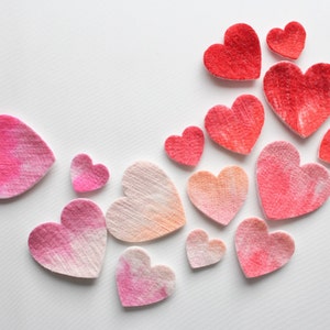 May include: A collection of felt hearts in various shades of pink and red, arranged in a curved pattern on a white background.