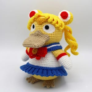 Puede incluir: Un peluche tejido a ganchillo que representa una criatura amarilla similar a un pato con pico color canela, vistiendo un traje de marinero blanco y azul con un lazo rojo. La figura tiene cabello largo amarillo en dos moños y pies palmeados.
