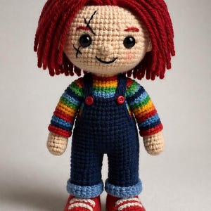 Könnte beinhalten: Handgemachte Häkelpuppe, die Chucky ähnelt. Die Puppe hat rote Garnhaare, aufgestickte Gesichtszüge und trägt einen regenbogenfarbenen gestreiften Pullover, eine blaue Latzhose und rote Schuhe. Die Puppe ist etwa 25 cm groß.