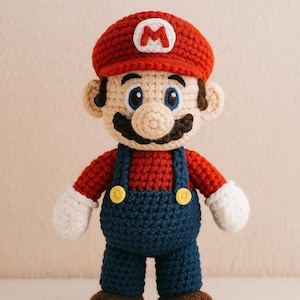 May include: A handmade crochet doll of the video game character Mario. The doll wears a red hat with a white "M", a red shirt, blue overalls, and brown shoes. It has a black mustache, blue eyes, and white gloves.