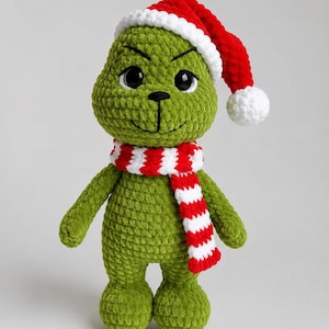 Peut inclure: Une poupée en peluche crochetée verte avec une surface en fil texturé, de grands yeux noirs, ainsi qu'un nez et une bouche brodés. La poupée porte un bonnet de Père Noël crocheté rouge et blanc avec un pompon blanc et une écharpe assortie.
