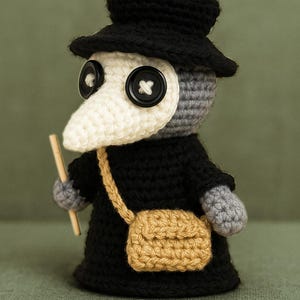 Puede incluir: Una muñeca de médico de la peste tejida a ganchillo con sombrero y túnica negros, que presenta una máscara de pico blanca con ojos de botón negros. La figura tiene manos grises, un bolso bandolera color canela y sostiene un palo de madera.