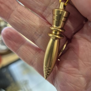 Op de afbeelding: Een gouden metalen slinger met een puntige punt en een decoratieve bovenkant. De slinger hangt aan een ketting.