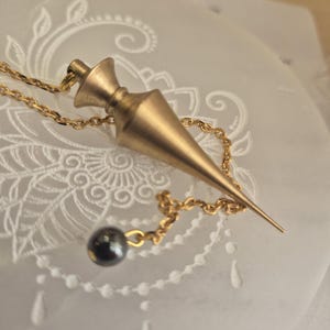 Könnte beinhalten: Ein goldfarbenes Metallpendel mit spitzer, konischer Form und ausgestelltem Oberteil, das an einer feinen goldfarbenen Kette hängt. Eine kleine, dunkle, kugelförmige Perle ist am Ende der Kette befestigt und liegt nahe am Pendelkörper.