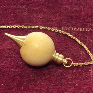 Könnte beinhalten: Ein Vintage-Pendel mit einem cremefarbenen, kugelförmigen Körper und einer goldfarbenen konischen Spitze. Das Pendel ist an einer goldfarbenen Kette befestigt. Der Hintergrund ist ein tiefrotes, samtiges Material. Das Wort "MONDIA" ist teilweise sichtbar.