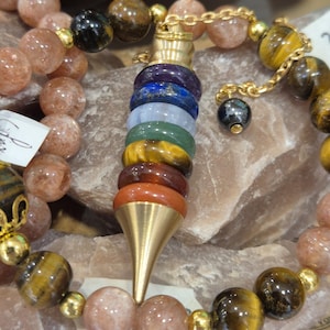 May include: A multi-colored chakra pendulum with a gold-colored cone-shaped tip and a gold chain, surrounded by beaded bracelets. The bracelets feature sunstone and tiger's eye beads, with gold-colored accents. The pendulum has a series of colored stones.