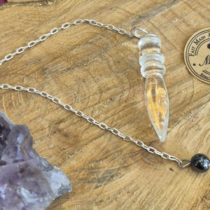 Könnte beinhalten: Ein klares Quarzkristall-Pendel mit einer silbernen Kette und einer schwarzen Hämatitperle. Das Pendel hängt an einer Holzoberfläche. "Fait Main Artisan Special" steht auf einem runden Etikett.