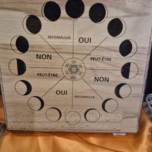 Può includere: Una tavola di legno quadrata con una disposizione circolare delle fasi lunari attorno a un design geometrico centrale. La tavola include etichette di testo in francese: OUI, NON, PEUT-ÊTRE e REFORMULER, organizzate in sezioni.