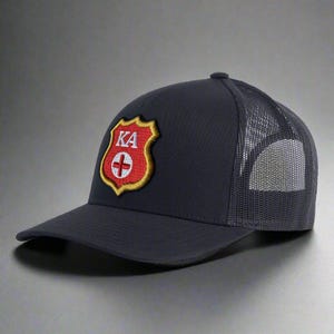 May include: Navy blue trucker hat with a mesh back and a curved brim. The front features a shield-shaped patch with the letters "KA" above a red cross. The patch is outlined in gold and red.
