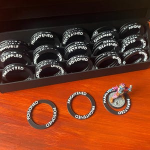 Könnte beinhalten: Eine schwarze Box mit schwarzen runden Token mit weißem Text, darunter Wörter wie "Frightened" und "Poisoned". Drei Token befinden sich außerhalb der Box, einer mit einer Miniaturfigur. Die Token sind für ein Tabletop-Spiel.