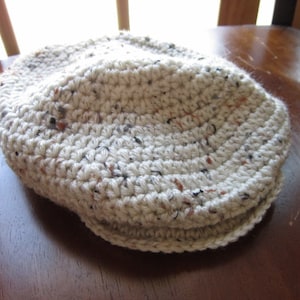 May include: A crocheted hat in a cream color with brown and black flecks. The hat has a rounded crown and a flat brim.