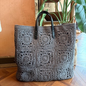 May include: A grey tote bag constructed from multiple crocheted granny squares featuring intricate circular patterns. The bag includes two black leather handles attached with metal rivets at the top, providing a structured contrast to the soft, textured grey yarn.
