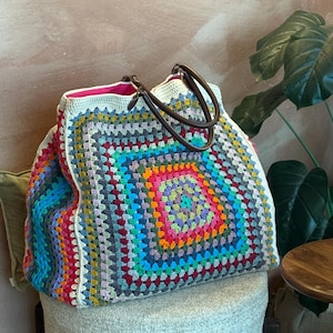 Puede incluir: Un bolso tote grande y colorido de ganchillo con un patrón cuadrado en una variedad de colores que incluyen rojo, naranja, amarillo, verde, azul y morado. El bolso tiene asas de cuero marrón y un forro interior rosa. El bolso está sobre un taburete claro.