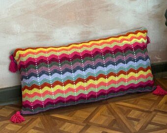 May include: A rectangular, handmade crochet pillow with a vibrant, zig-zag pattern in shades of yellow, pink, purple, and green. The pillow has pink tassels on each end. This decorative pillow adds a pop of color to any room.