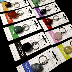 May include: Keychains featuring various gemstones, each attached to a silver-colored keyring. Each keychain is displayed on a white card with a colored banner indicating a zodiac sign. The gemstones vary in color and texture.