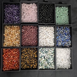 May include: A selection of gemstone beads displayed in small black boxes. The beads are in a variety of colours, such as purple, pink, black, green, yellow, red, brown, white, blue, and clear. The beads are round and polished.