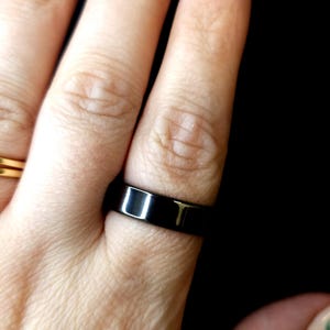 Puede incluir: Un anillo negro de forma rectangular se lleva en un dedo. El anillo tiene una superficie brillante y reflectante. El fondo es negro.