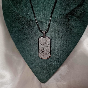 Può includere: Una collana con un ciondolo a forma di targhetta per cani in argento su un cordoncino nero. La targhetta è realizzata in materiale meteoritico e presenta un motivo unico. Il numero "1" è visibile sulla targhetta.